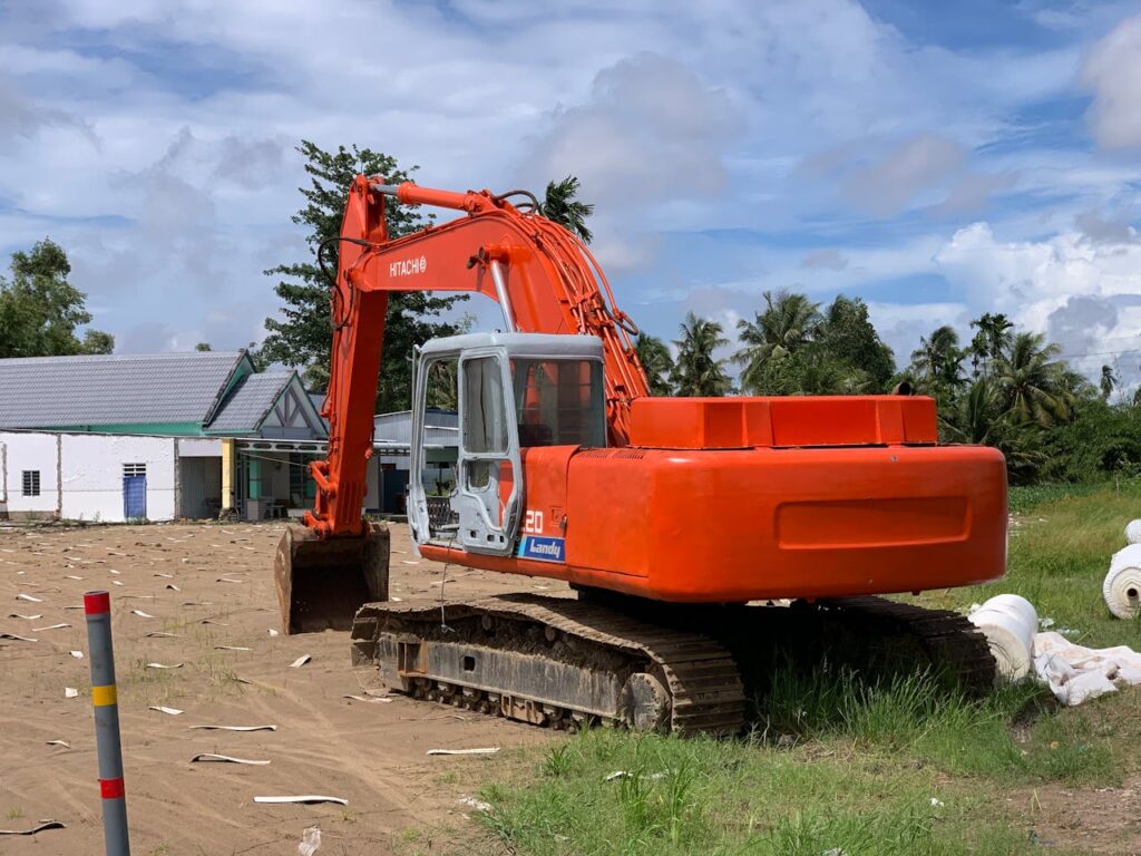 An orange excavator stands idle at a sunny construction site with tropical greenery.